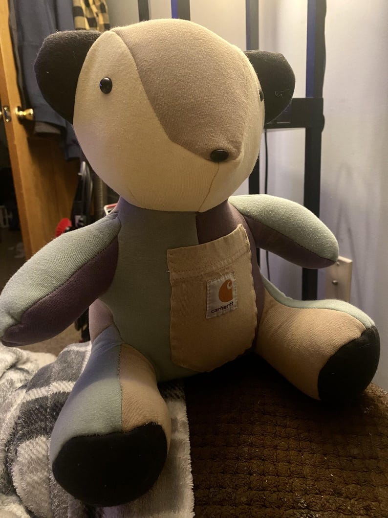 May include: A patchwork teddy bear in various shades of beige, grey, and blue. The bear has black button eyes and a small pocket with the Carhartt logo. The bear is a handmade item.