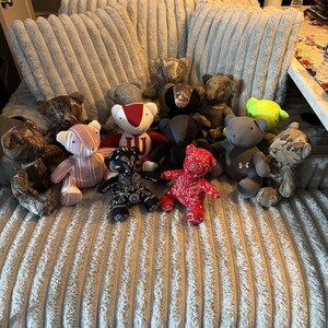 May include: A group of teddy bears in diverse colours and patterns, such as camouflage, red paisley, and stripes. Some bears feature sports team logos. The bears are displayed on a textured, light grey surface.