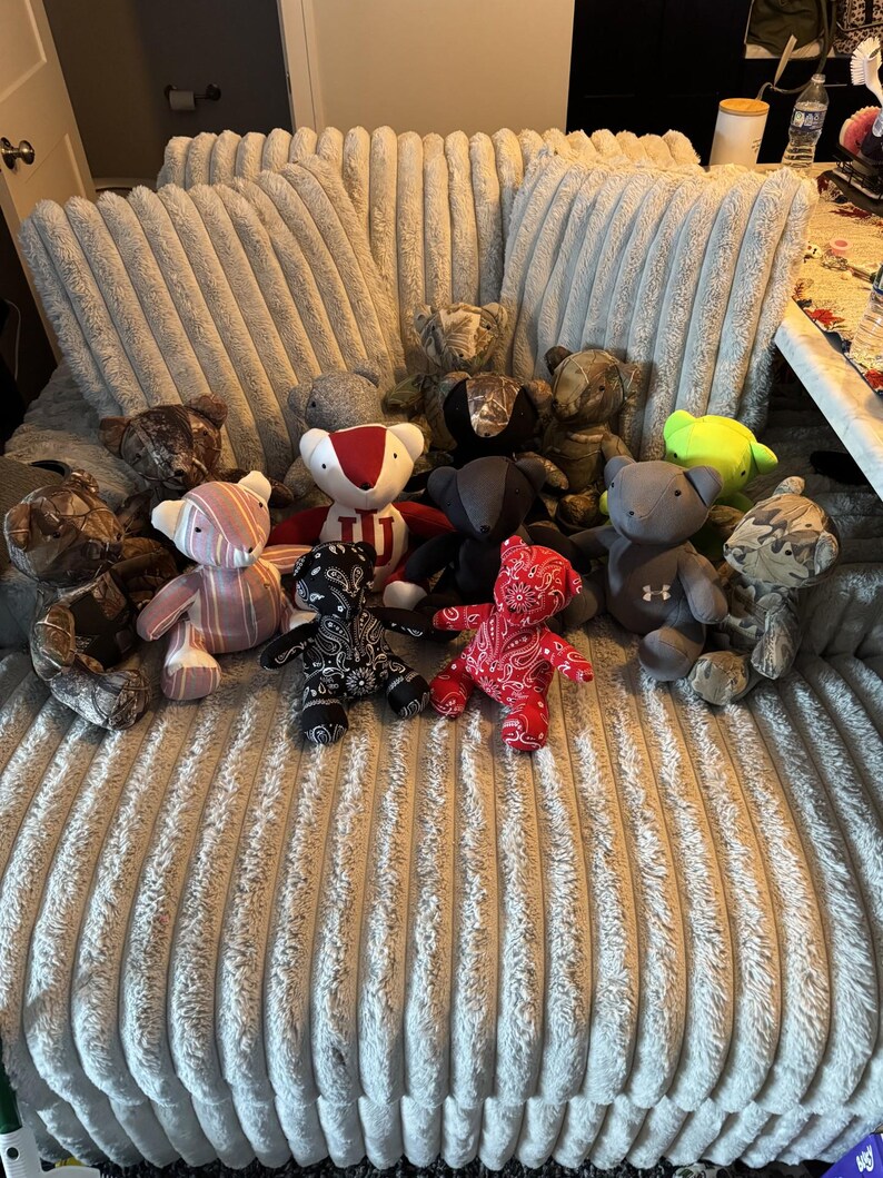 May include: A group of teddy bears in diverse colours and patterns, such as camouflage, red paisley, and stripes. Some bears feature sports team logos. The bears are displayed on a textured, light grey surface.