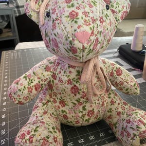 May include: A handmade teddy bear with a floral pattern in pink and red on a cream background. The bear has a pink heart-shaped nose, black eyes, and a pink bow. The bear is sitting on a grey cutting mat.