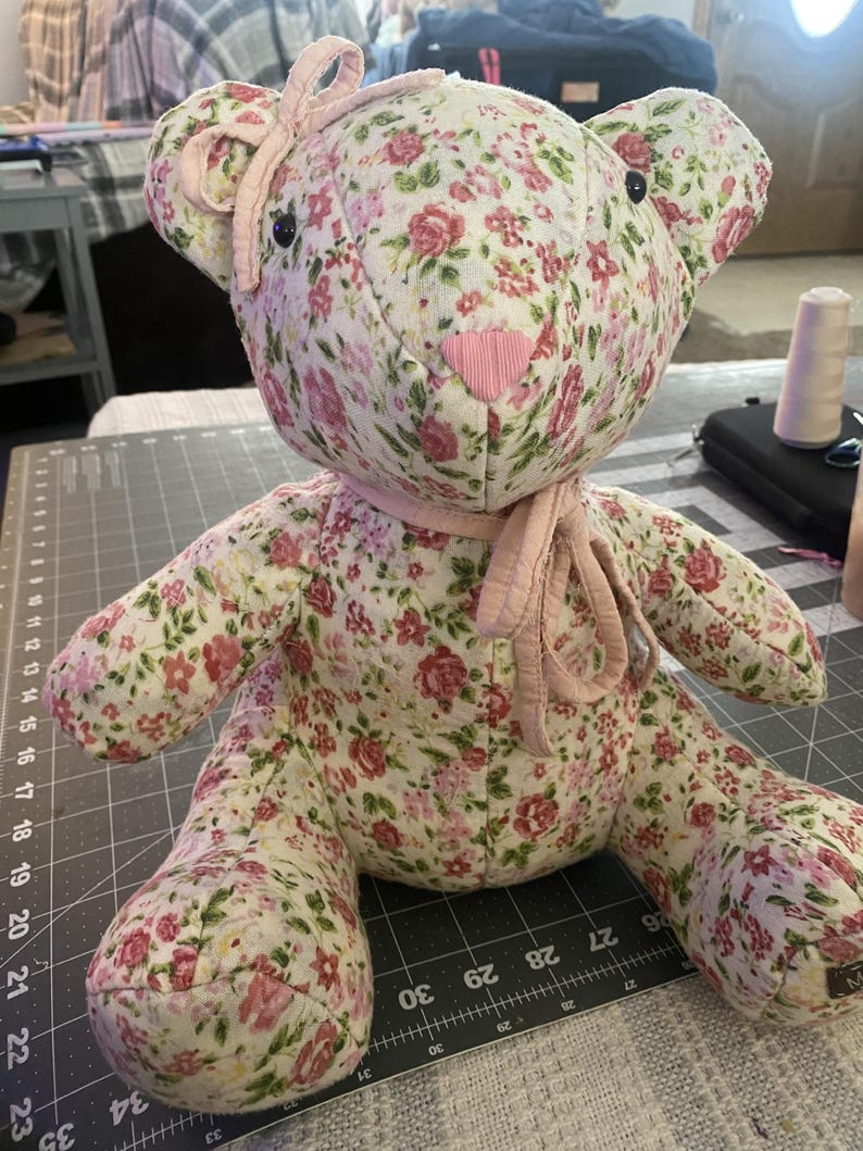 May include: A handmade teddy bear with a floral pattern in pink and red on a cream background. The bear has a pink heart-shaped nose, black eyes, and a pink bow. The bear is sitting on a grey cutting mat.