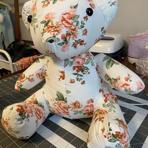May include: A handmade teddy bear with a floral pattern. The bear is made of white fabric with a pattern of pink and orange roses and green leaves. The bear's ears are made of white lace. The bear has black button eyes and a black stitched nose.