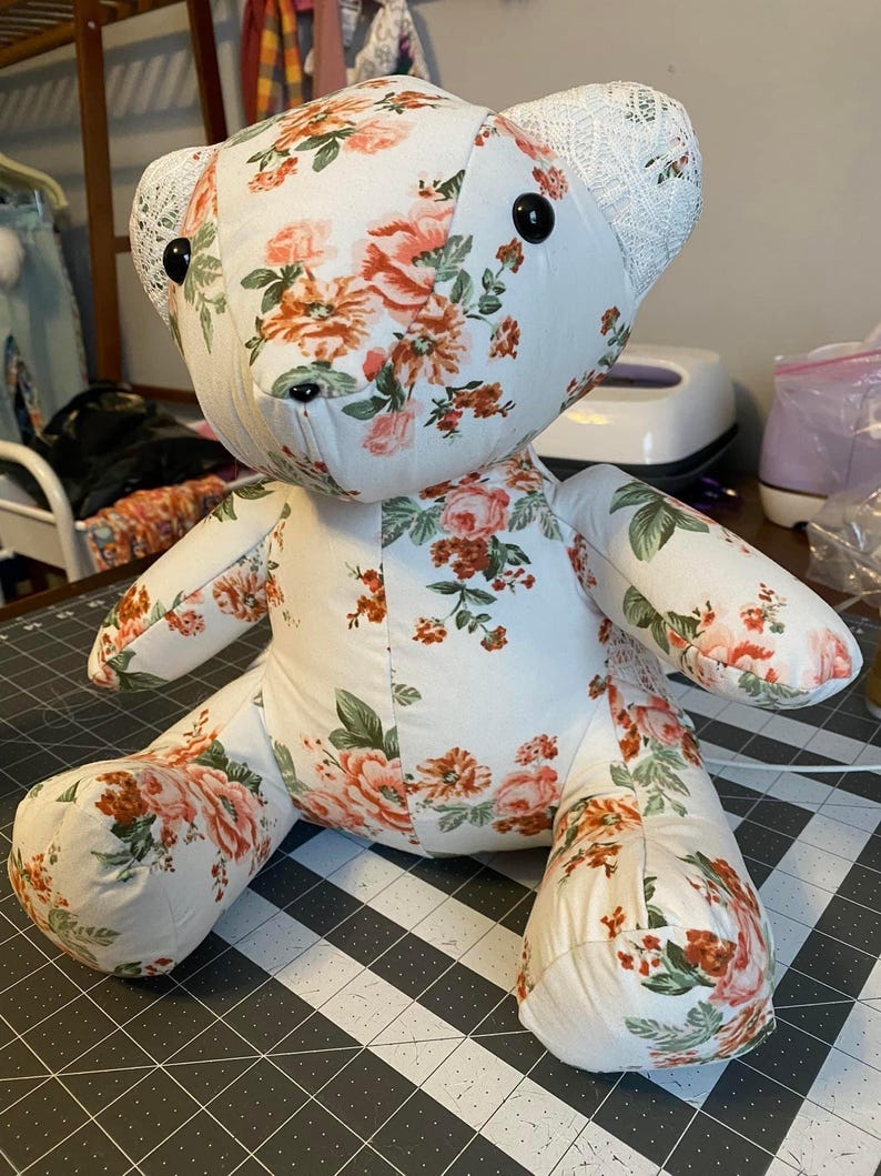 May include: A handmade teddy bear with a floral pattern. The bear is made of white fabric with a pattern of pink and orange roses and green leaves. The bear's ears are made of white lace. The bear has black button eyes and a black stitched nose.