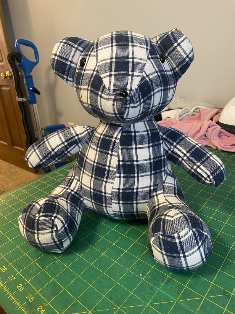 May include: A handmade teddy bear made from navy blue and white plaid fabric. The bear has black button eyes and is sitting on a green cutting mat. The bear's arms and legs are outstretched.