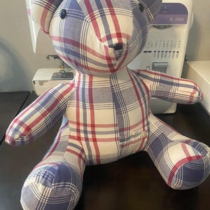 May include: A plaid teddy bear in red, white, and blue. The bear has black button eyes and a small button on its chest. The fabric appears to be cotton or a similar material. The bear is sitting upright. The plaid pattern is a classic design.