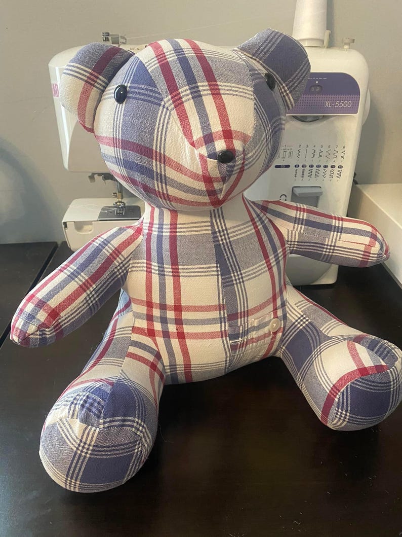 May include: A plaid teddy bear in red, white, and blue. The bear has black button eyes and a small button on its chest. The fabric appears to be cotton or a similar material. The bear is sitting upright. The plaid pattern is a classic design.