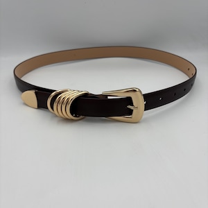 Op de afbeelding: Een donkerbruine riem met een goudkleurige gesp en decoratieve ringen. De riem is gemaakt van een glad materiaal en heeft een klassiek ontwerp. De riem is ongeveer 2,5 cm breed.