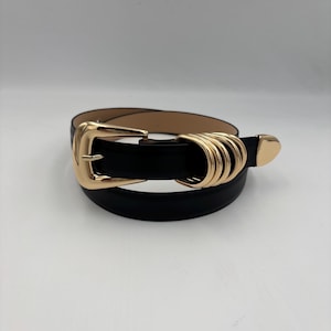 May include: Black belt with a gold-coloured buckle and decorative gold-coloured loops. The belt has a classic design with a rectangular buckle and a pointed end. The belt is likely made of leather or a leather-like material.