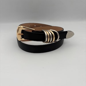May include: Black belt with a gold buckle and silver and gold ring accents. The belt has a pointed silver tip. The belt is made of a smooth, black material.