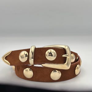 Op de afbeelding: Een bruine suède riem met goudkleurige metalen studs en een gesp. De riem is dubbelgevouwen en de gesp is zichtbaar.