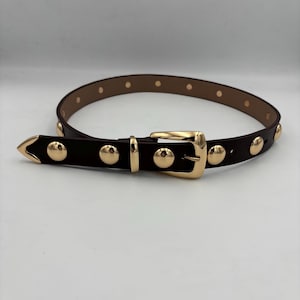 Peut inclure: Une ceinture marron foncé avec des clous dorés et une boucle dorée. La ceinture a une extrémité pointue et est ornée d'une série de clous arrondis espacés. La boucle est rectangulaire.