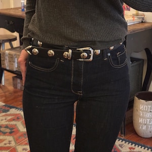May include: A black leather belt with silver studs and a silver buckle. The belt is worn over a pair of dark blue jeans.