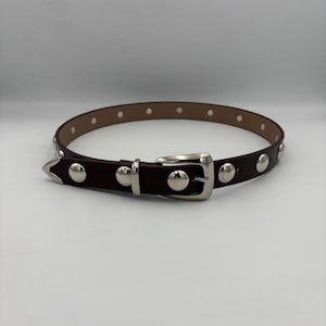 May include: A brown leather belt with silver studs and a silver buckle. The belt features a classic design with a pointed end and evenly spaced studs. The rectangular buckle has a polished finish. The belt is approximately 100 cm in length.