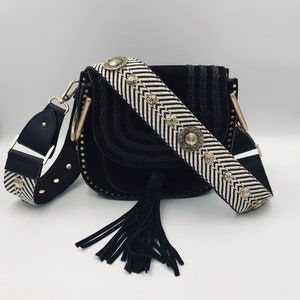 Peut inclure: Sac à bandoulière en daim noir avec une bandoulière rayée noir et blanc avec des accents dorés. Le sac a un pompon et un détail tressé.