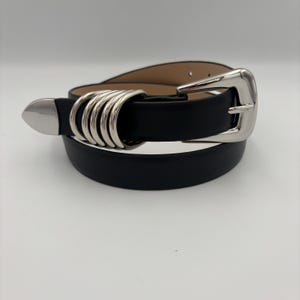Op de afbeelding: Een zwarte leren riem met een zilveren gesp met een ringontwerp. De gesp is in een eenvoudige, klassieke stijl.