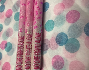 Könnte beinhalten: Drei rosa Glitzer-Stifte mit silbernen Akzenten. Jeder Stift trägt den Schriftzug "No one fights alone" in rosa. Die Stifte befinden sich auf einem rosa-blauen Punkte-Hintergrund.
