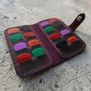 May include: Open leather guitar pick holder with various colored picks. The holder is dark brown with purple stitching and a snap closure. The picks are in various colors, including red, green, purple, and orange.