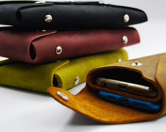 May include: Leather phone cases in black, burgundy, and olive green are stacked, with a fourth case open to reveal two smartphones. The cases have a snap closure and are made of textured leather.