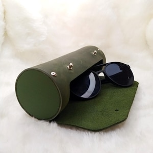 May include: A green leather cylindrical sunglasses case with a flap that opens to reveal a pair of black sunglasses.