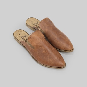 Pode incluir: Um par de mules de couro castanho. Os sapatos têm bico fino e acabamento desgastado. As solas são castanho claro com uma borda costurada. Os sapatos são exibidos em um fundo cinza claro.