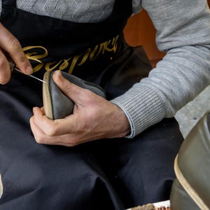 A man wearing a gray sweater and a black apron is carefully crafting a pair of shoes, using a tool to shape and adjust the leather material.