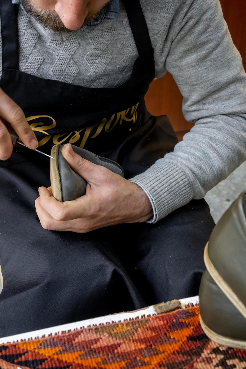 A man wearing a gray sweater and a black apron is carefully crafting a pair of shoes, using a tool to shape and adjust the leather material.