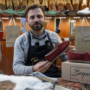 A man in a gray shirt and apron is holding up a red shoe in a shoe store, surrounded by various shoes displayed on shelves.