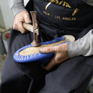 A person wearing a black apron with gold text is working on a blue object, which appears to be a piece of footwear.