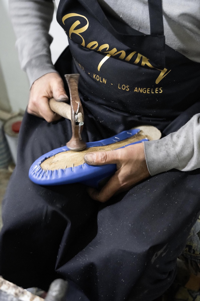 A person wearing a black apron with gold text is working on a blue object, which appears to be a piece of footwear.