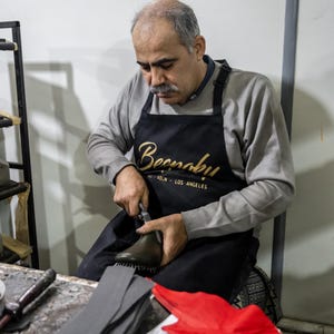 A man wearing a black apron with the word "Beenah" is working on a pair of shoes, holding a tool in his hand.