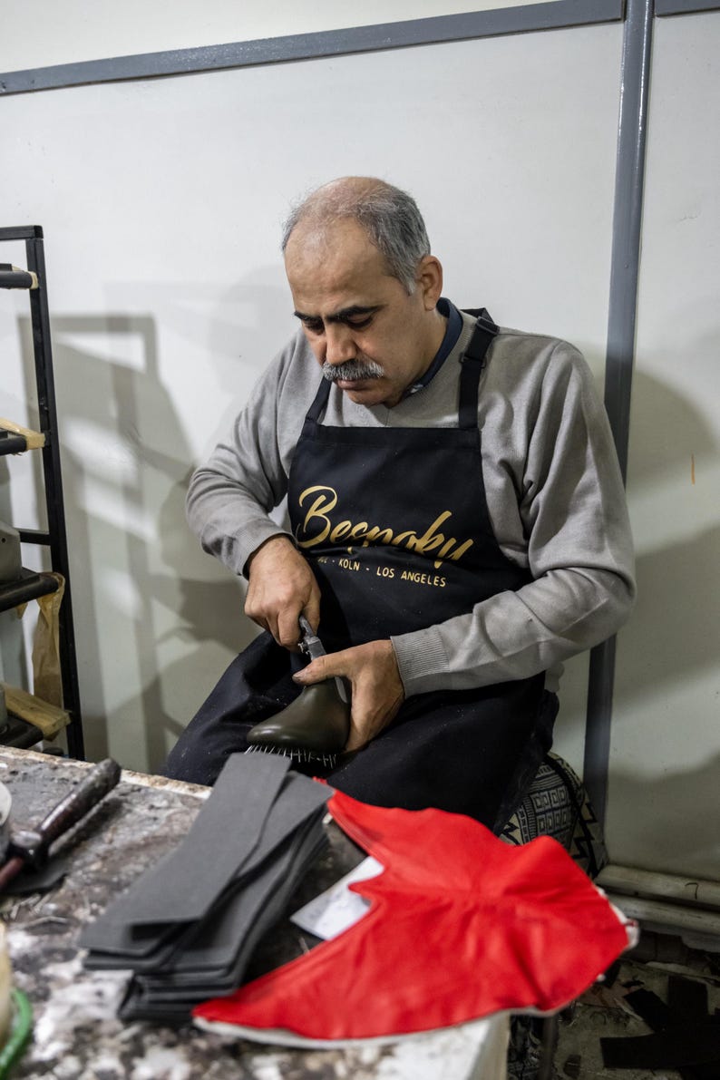 A man wearing a black apron with the word "Beenah" is working on a pair of shoes, holding a tool in his hand.