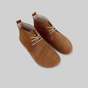 May include: A pair of brown suede ankle boots with white laces. The boots have a rounded toe and a pull tab at the back. The shoes are displayed on a light gray background.