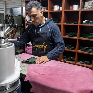 A man in a blue sweatshirt and apron is working on a pink leather item in a workshop, using a machine to shape the leather.