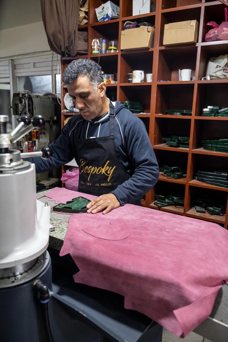 A man in a blue sweatshirt and apron is working on a pink leather item in a workshop, using a machine to shape the leather.