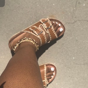 May include: Brown leather sandals with multiple straps and buckles. The sandals have a white stitched trim and a white sole. The sandals are worn on a person's feet.