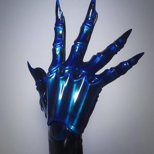 May include: A blue, claw-like glove with long, pointed fingernails. The glove has a metallic sheen, reflecting light with shades of blue and teal. The design features articulated segments, giving the appearance of a skeletal hand.