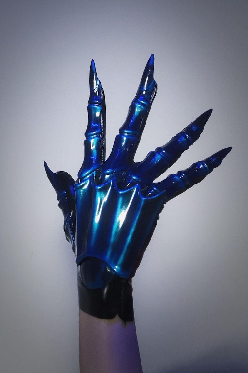 May include: A blue, claw-like glove with long, pointed fingernails. The glove has a metallic sheen, reflecting light with shades of blue and teal. The design features articulated segments, giving the appearance of a skeletal hand.