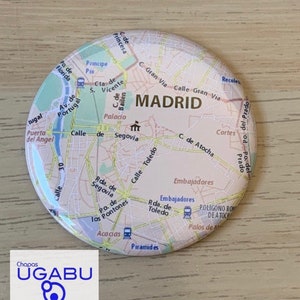 Puede incluir: Un botón redondo con un mapa de Madrid, España, con nombres de calles y puntos de referencia. El botón tiene un acabado brillante y un borde blanco. El texto "MADRID" es claramente visible. El botón está sobre una superficie de madera clara.
