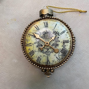 May include: A gold-colored ornament shaped like a pocket watch with Roman numeral markings and a detailed design on the face. The ornament has a gold-colored string attached to the top.