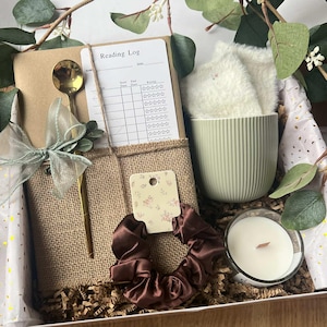 May include: A gift box containing a brown notebook with a burlap band, a gold spoon, a reading log card, a dark brown satin scrunchie, a sage green ribbed mug with white fuzzy socks, and a small candle in a glass jar, decorated with eucalyptus.