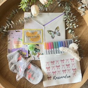 May include: A gift set containing a Soou blueberry vitamin-C mask, a 24K gold eye patch packet, a small green container, a set of colourful markers, a pair of grey fuzzy socks with pink flamingos, and a white pouch with pink bows and the name Kassandra.