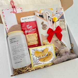 Puede incluir: Una caja de regalo que contiene un vaso de vidrio esmerilado con texto para amantes de los libros, un paquete de capuchino de vainilla francesa Tim Hortons, una mascarilla de ojos 24K gold, un lazo rojo, un bloc de notas con cerezas, un colgante dorado y una pinza de pelo gris.