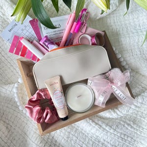 May include: A wooden tray filled with self-care items. Includes a white cosmetic bag, pink pens, a candle, a scrunchie, a tube of hand cream, and a face mask. The tray is decorated with a white beaded garland and a pink ribbon that says "Just For You".