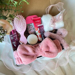 Può includere: Un cofanetto regalo rosa pieno di prodotti di bellezza. Include una spazzola per capelli rosa, una fascia per capelli e vari articoli per la cura della pelle come crema per le mani e balsamo per le labbra. La scatola è legata con un nastro rosa.