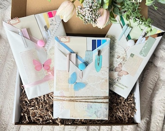 May include: Three rectangular stationery sets wrapped in white paper with vintage-style collage prints, butterfly embellishments, and metal clips. Each set includes pens, paper inserts, and decorative accents, all arranged inside a brown cardboard box.
