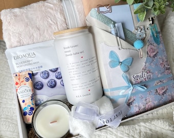 May include: A gift box containing a white glass tumbler with book-lover text, a blueberry moisturizing mask, a Sadoer hand cream, a white candle, a floral notebook with a blue butterfly clip, a pen, and a white fuzzy blanket for cozy reading.