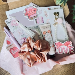 May include: A gift box containing a floral bookmark with a white tassel, a light purple pen, a peach-colored satin scrunchie, a tube of Sadoer rose hand cream, a pearl-beaded keychain with a pink pom-pom, and stickers featuring the text "Fell in love on the third chapter" and "Love" with a pink bow.
