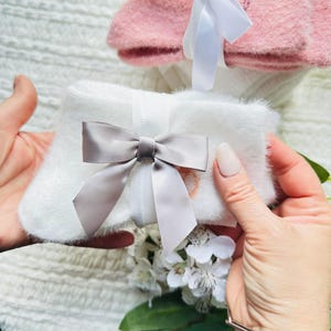 May include: A pair of white fluffy socks tied with a gray satin bow. A white ribbon is also tied around the socks. A pair of pink socks is visible in the background. The socks are presented on a white textured surface.