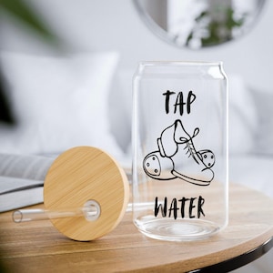 Pode incluir: Um copo transparente em forma de lata com tampa de bambu e palhinha de vidro. O copo apresenta um gráfico preto de um sapato de sapateado e o texto "TAP WATER" impresso numa fonte de estilo manuscrito e negrito na parte frontal.