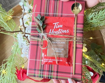 Op de afbeelding: Een cadeau verpakt in rood ruitpatroon, vastgebonden met touw en versierd met groen. Een zakje Tim Hortons warme chocolademelk en een gouden lepel zijn inbegrepen. Het cadeau wordt door een persoon vastgehouden.